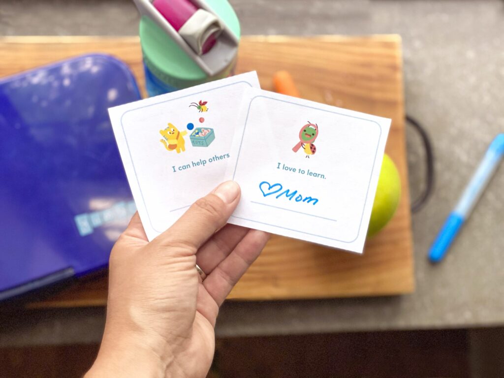 Free printable lunchbox notes held in a parent's hand over a lunchbox, apple, and water bottle